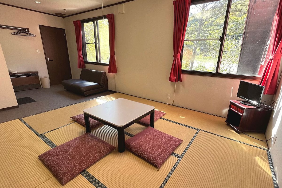A room with tatami flooring, a sofa, a table, a TV, and a window with red curtains.