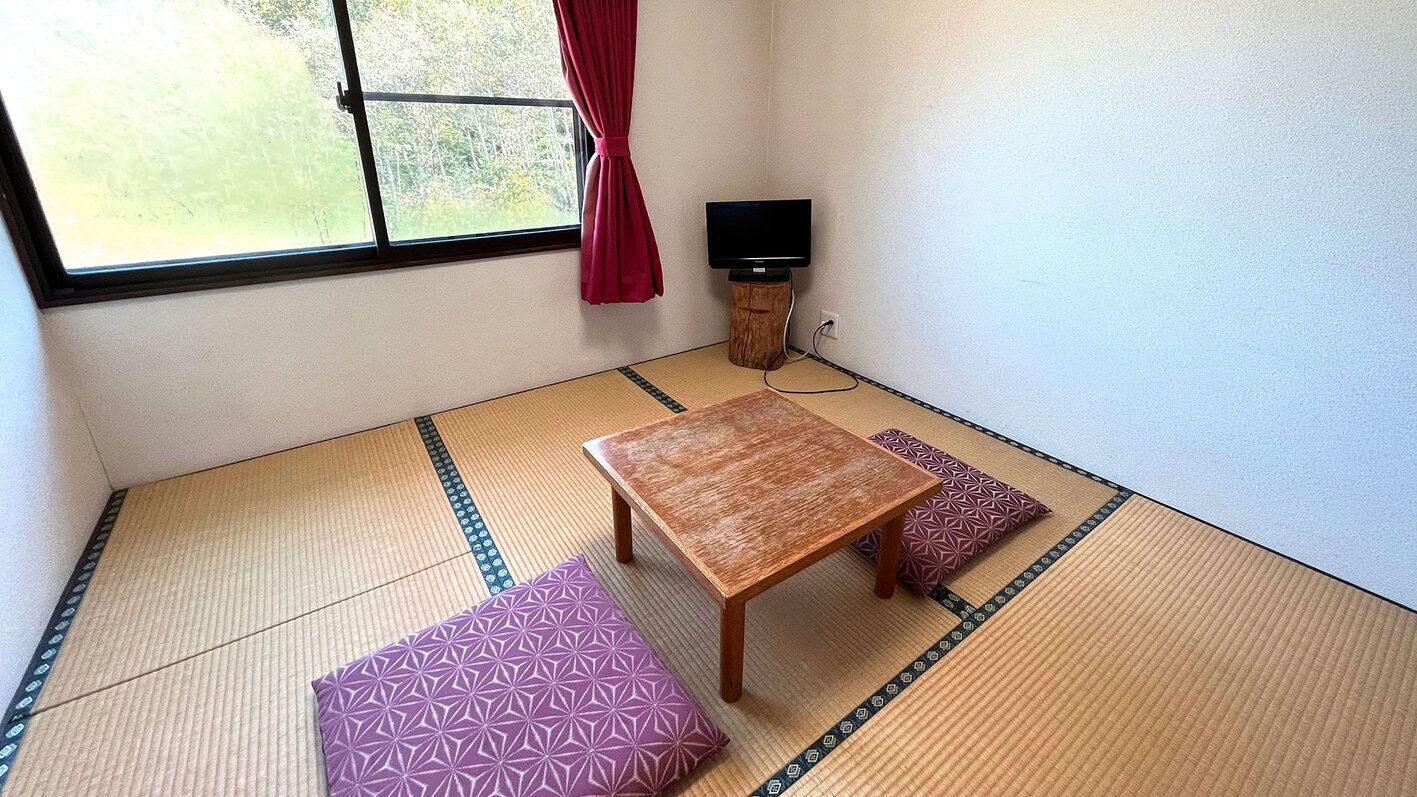 Ein Zimmer mit Tatami-Matten, einem Holztisch und Kissen, einem Fernseher auf einem Holzständer sowie einem Fenster mit Vorhang.