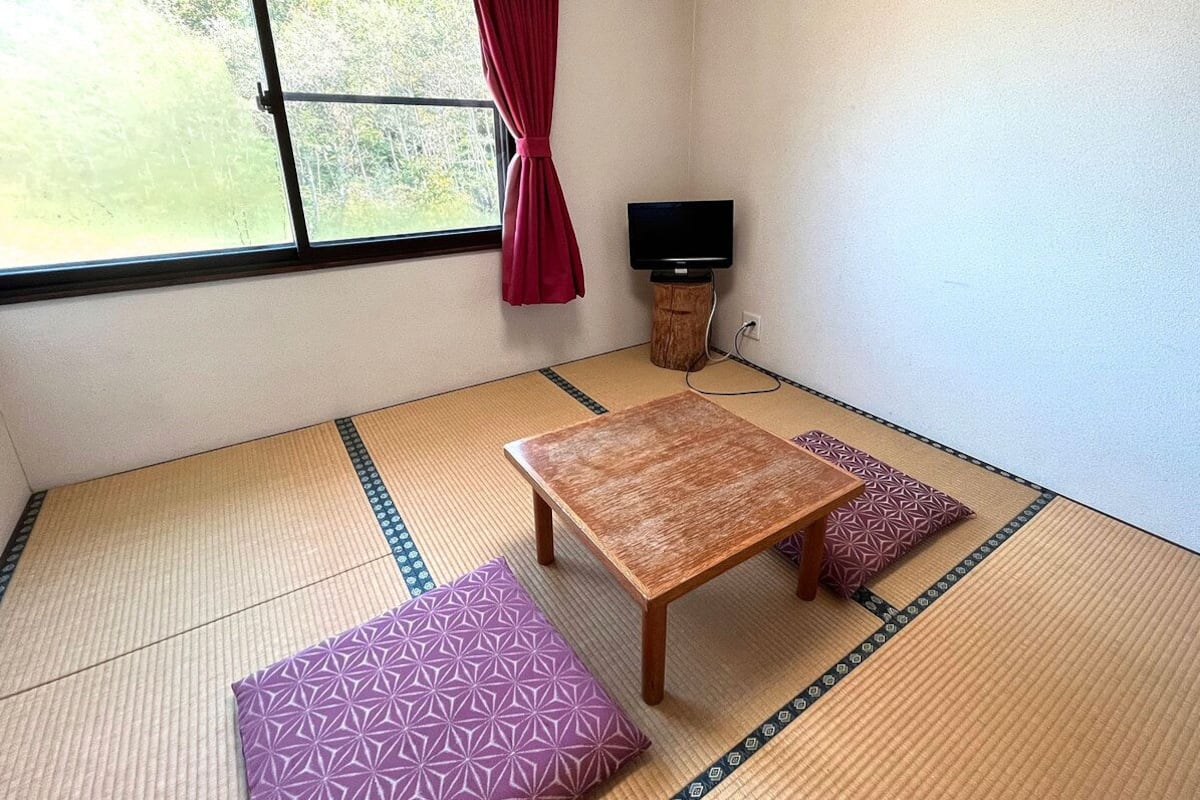 A tatami mat room with a wooden table and cushions, a TV on a wooden stand, and a window with a curtain.
