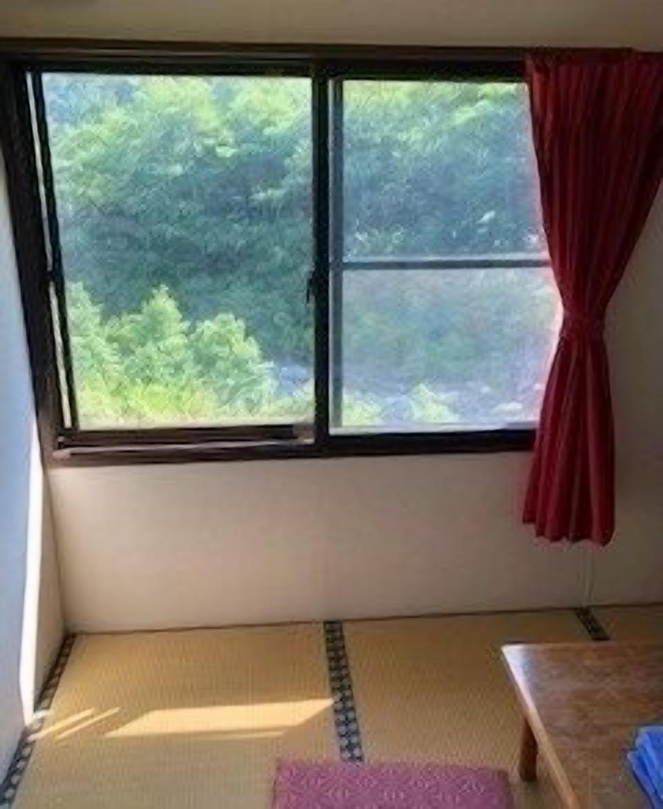 A room with tatami flooring, a window with a view of greenery, and a red curtain.