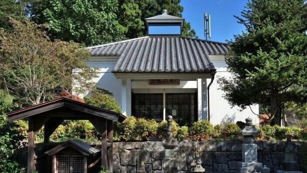 Un bâtiment traditionnel japonais, avec un toit recouvert de tuiles, un mur de pierre et une petite structure en bois au premier plan.
