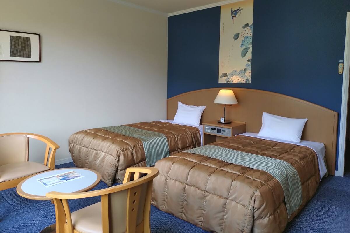 Habitación de hotel con dos camas, una mesita pequeña, una silla y una mesita de noche fijada a la pared.