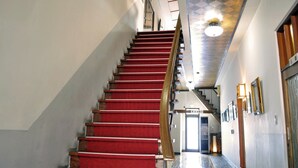 Una escalera con alfombra roja que lleva a una plataforma, un techo con diseño geométrico y un pasillo con cuadros en la pared.