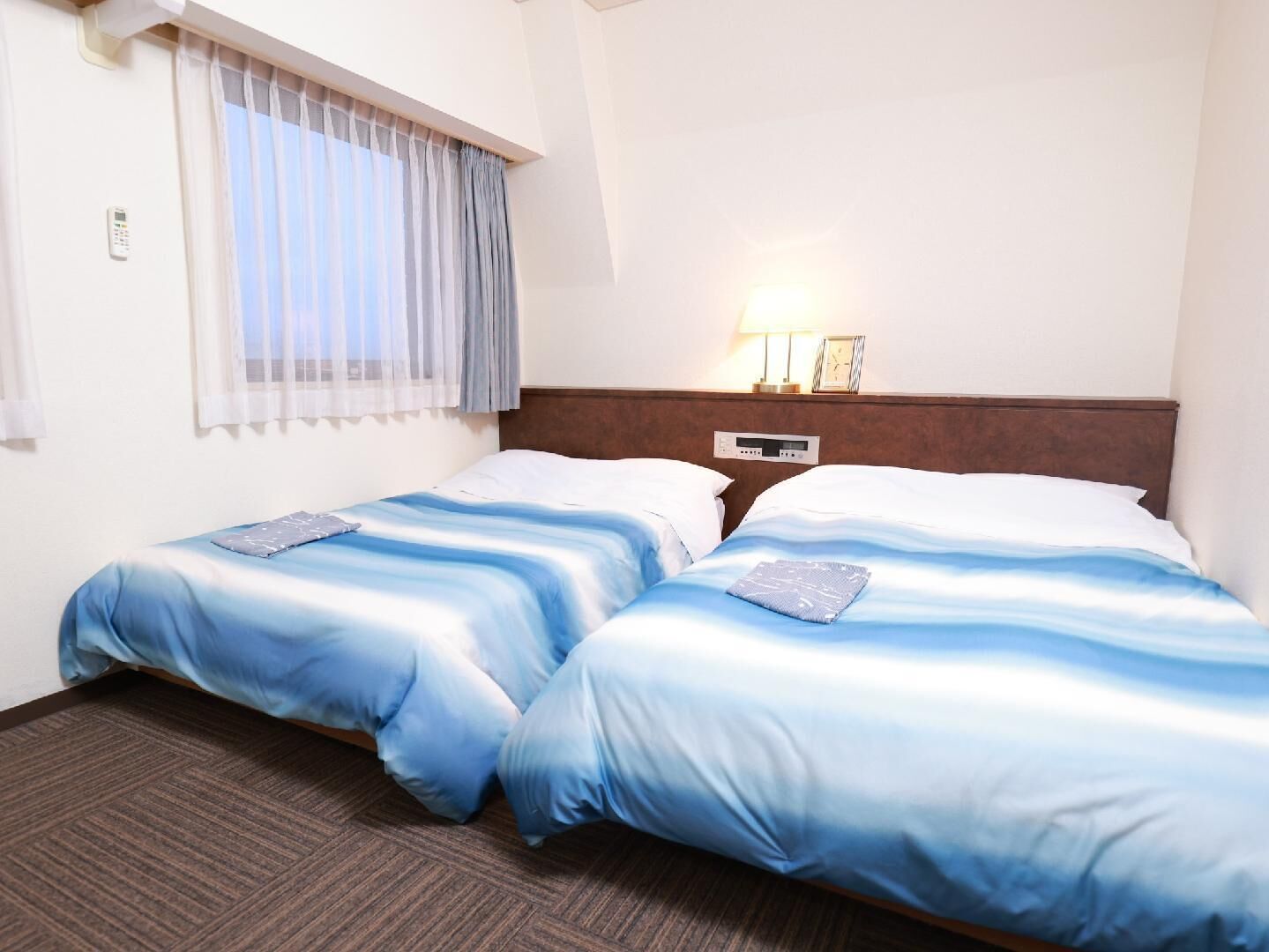 Hotellihuone, jossa on kaksi sänkyä, ikkuna, jossa on verhot, yöpöytä lampun kanssa ja seinäkello.