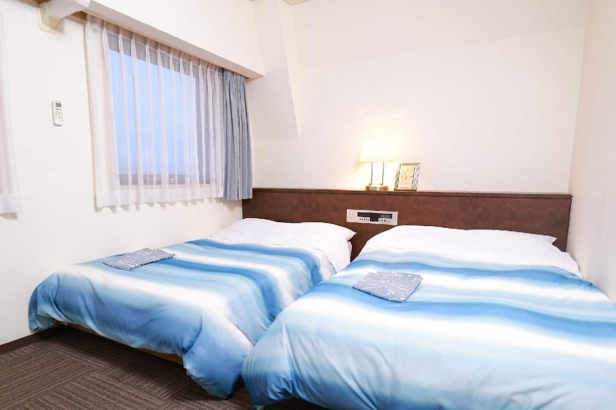 Habitación de hotel con dos camas, una ventana con cortinas, una mesita de noche con lámpara y un reloj de pared.