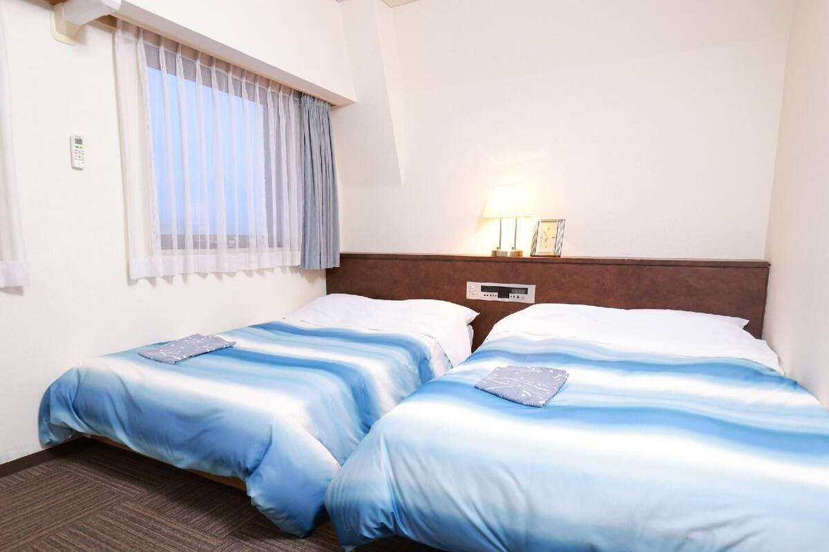 Hotellihuone, jossa on kaksi sänkyä, ikkuna, jossa on verhot, yöpöytä lampun kanssa ja seinäkello.