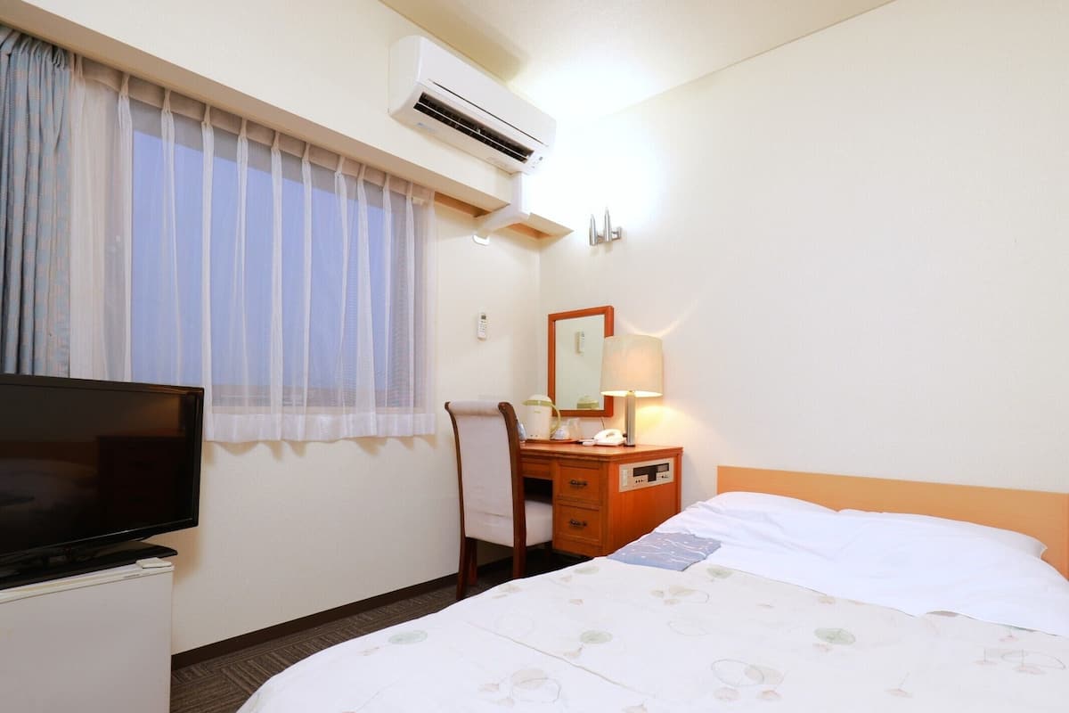 Una habitación de hotel con cama, televisión, escritorio con lámpara, espejo y aire acondicionado.