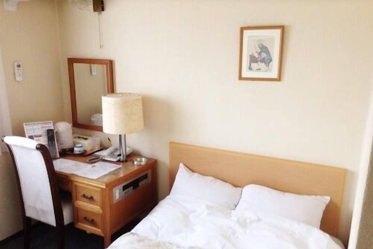 Habitación de hotel con cama, escritorio con lámpara, silla, espejo y un cuadro en la pared.