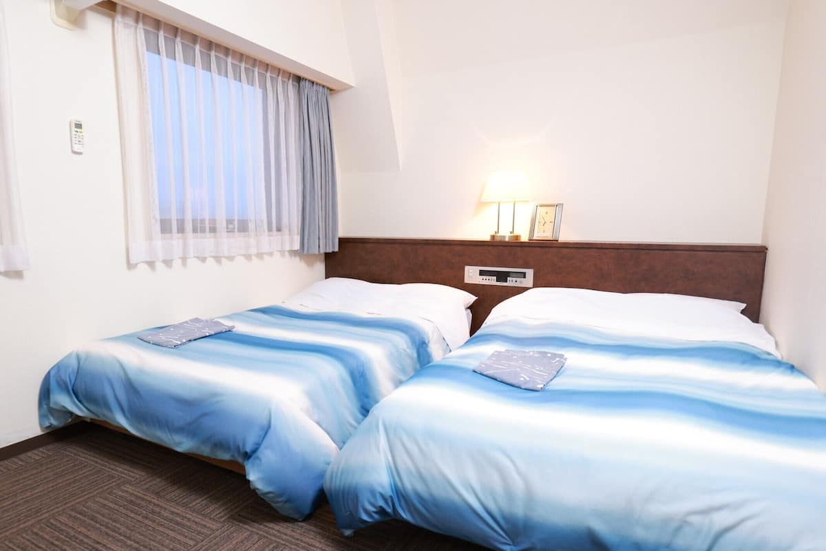 Habitación de hotel con dos camas, una mesita de noche, un reloj y una ventana con cortinas.
