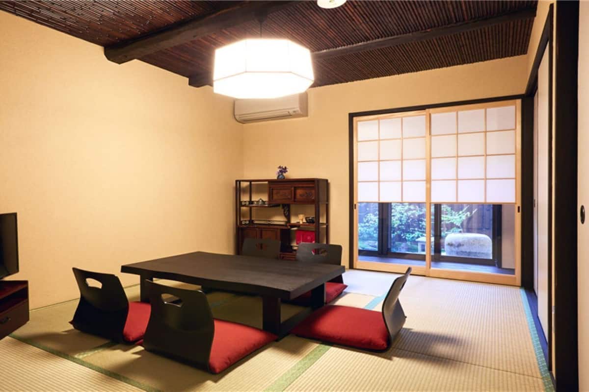 Ein traditioneller japanischer Raum mit Tatami-Boden, einem niedrigen Tisch, Kissen, einem Fernseher und einer Schiebetür mit Blick.