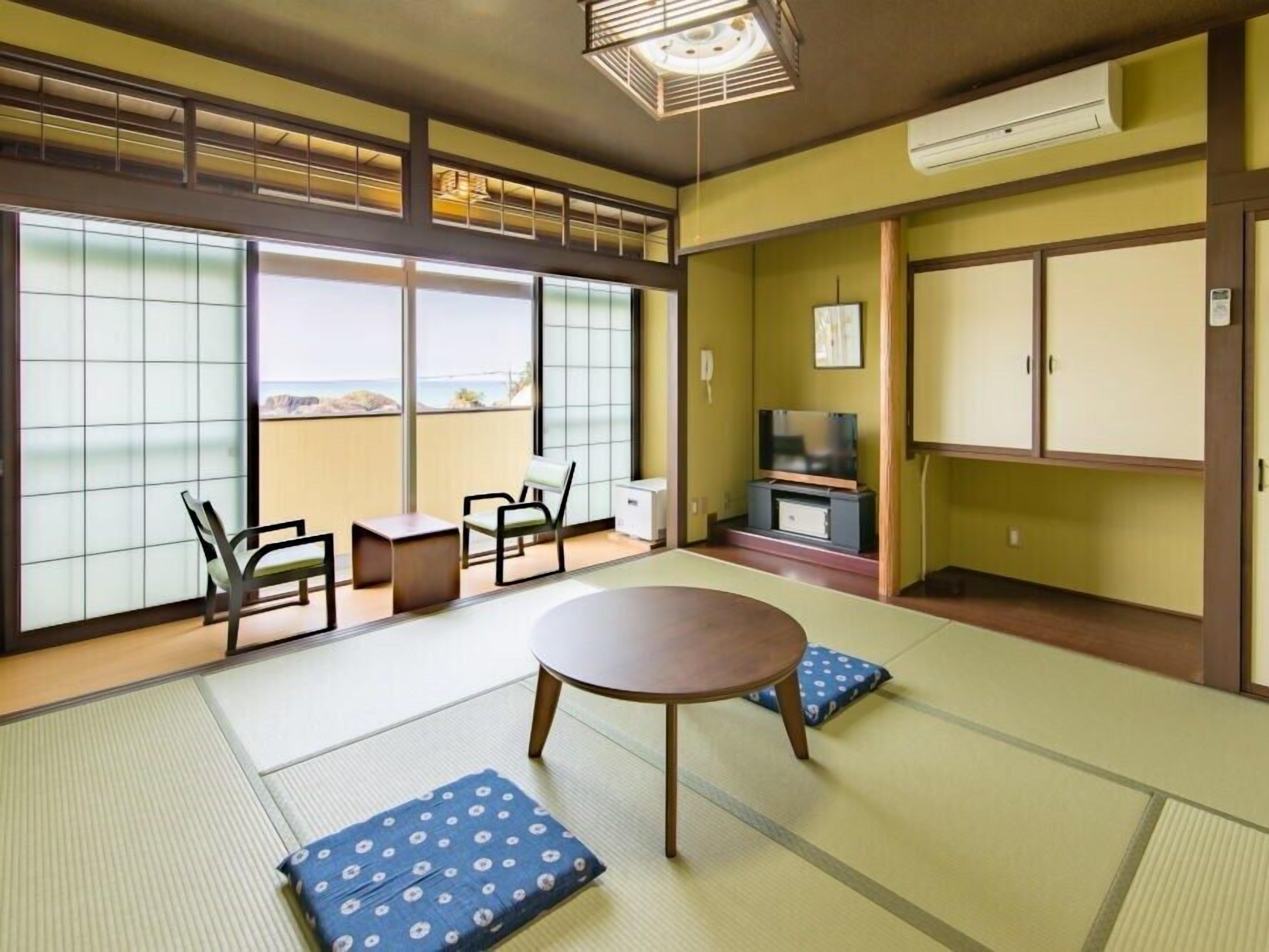 A traditional Japanese room with tatami flooring, a low table, chairs, a television, and a view of the sea.