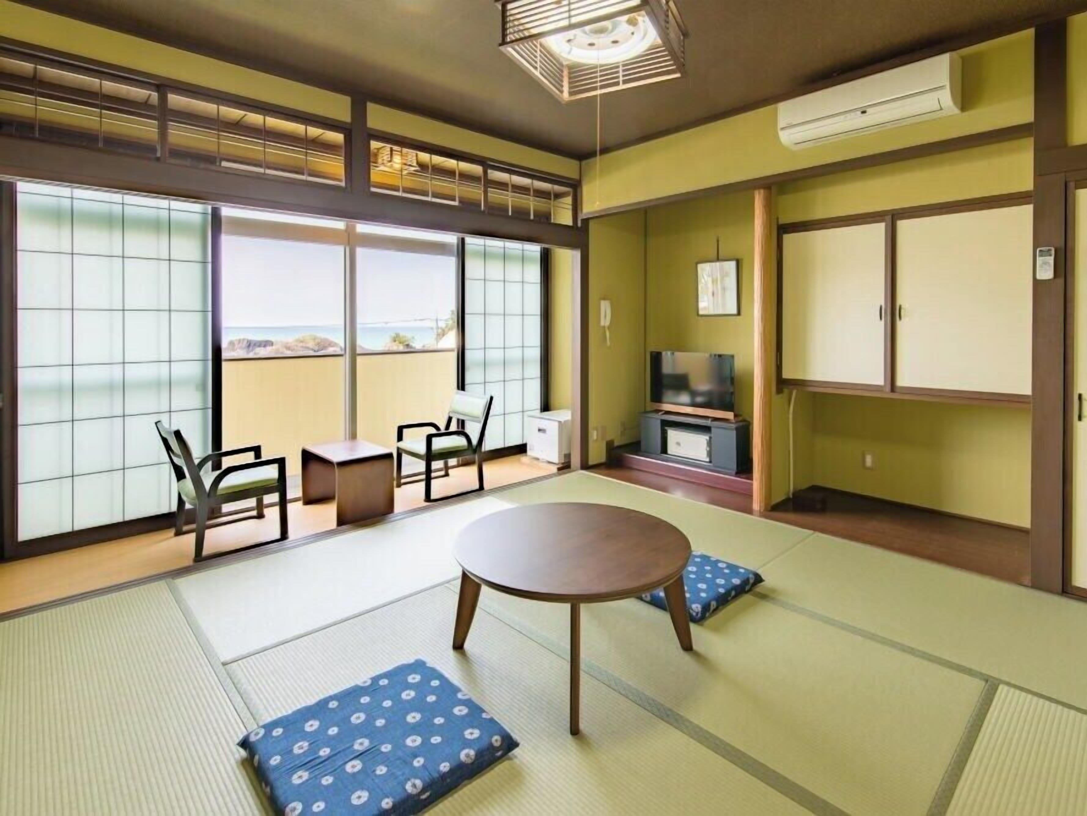 A room with tatami mats, a low table, chairs, a TV, and a view of the sea.