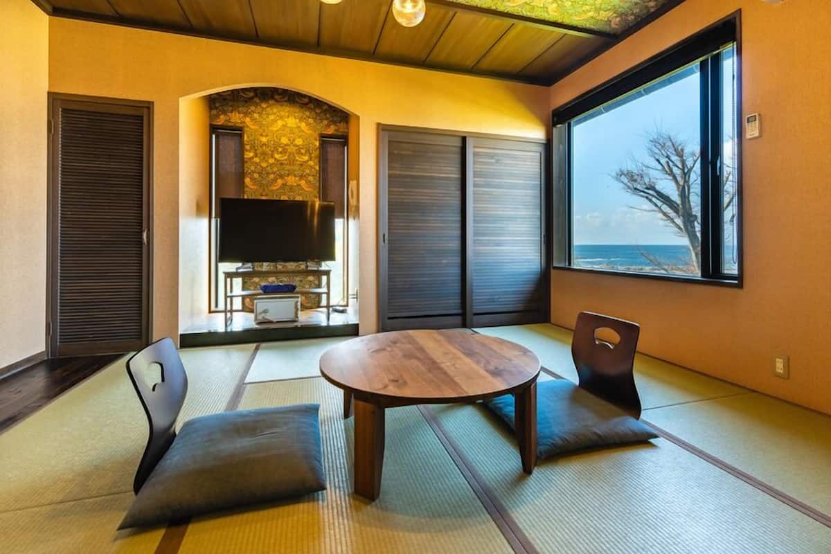 Ein Zimmer mit Tatami-Matten, einem runden Tisch, Stühlen, einem Fernseher, Schiebetüren und Blick auf das Meer.