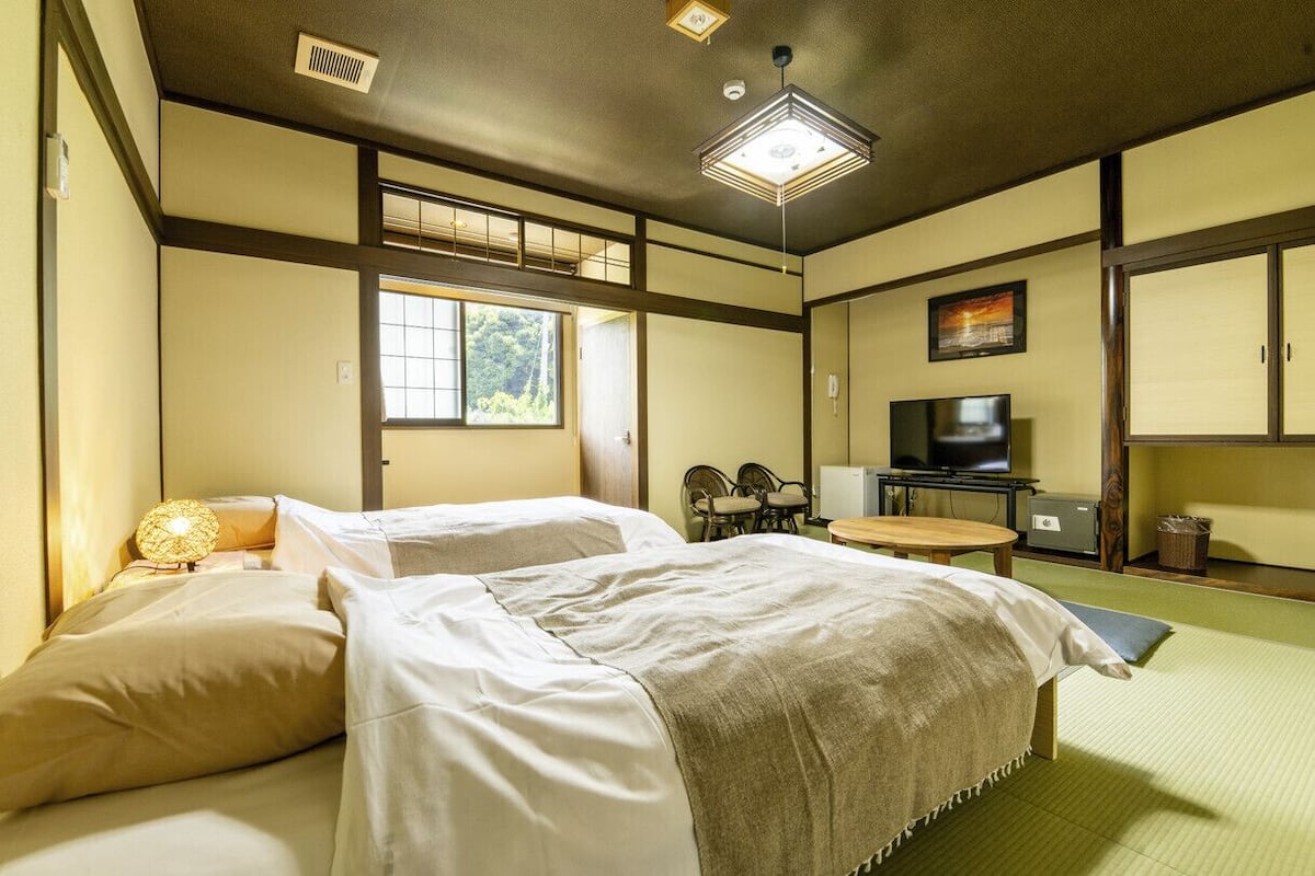 Ein traditioneller japanischer Raum mit Tatami-Boden, einem Bett mit weißer Bettwäsche, einem Fernseher und einem Schreibtisch mit Stühlen.