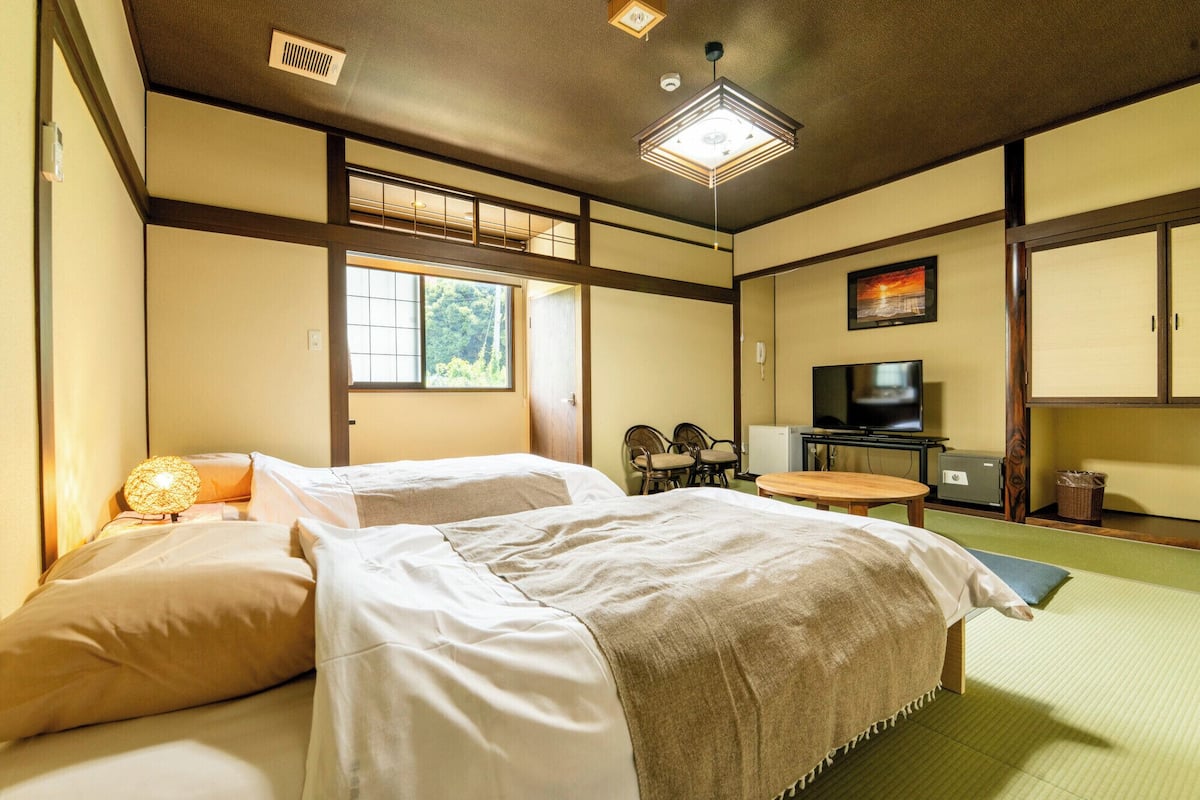 Ein traditioneller japanischer Raum mit Tatami-Böden, zwei Betten, einem Fernseher und einem Fenster mit Blick auf Grünflächen.