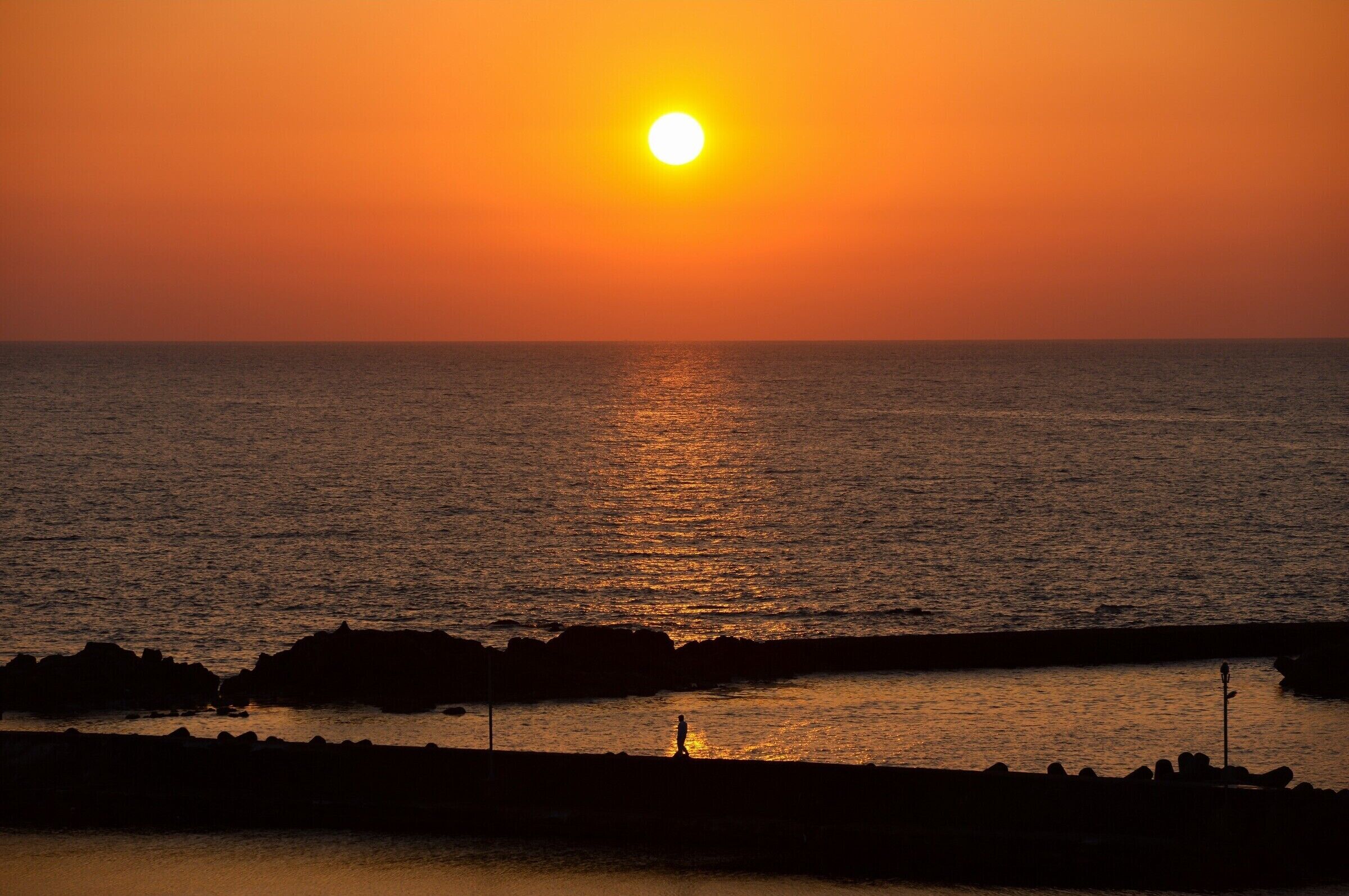 A sunset over the ocean with a silhouette of a person walking along a rocky shoreline.