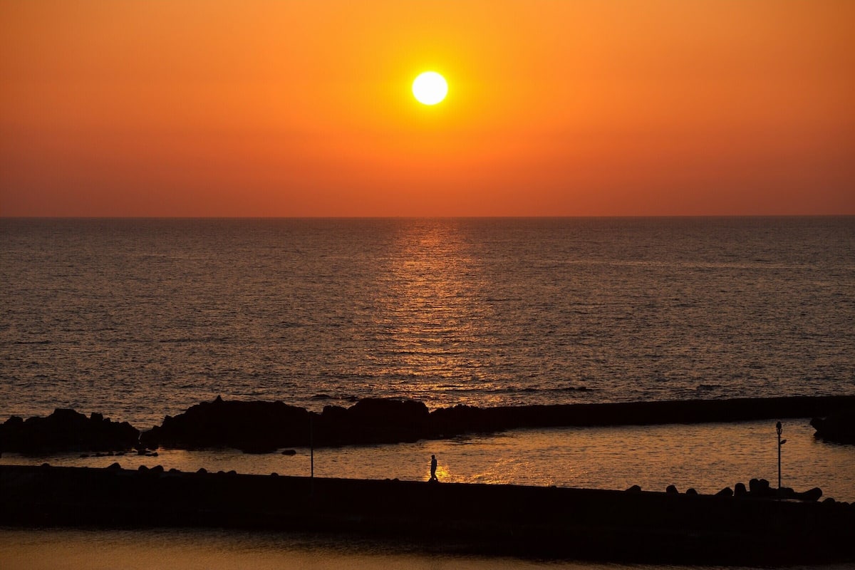 Ein Sonnenuntergang über dem Meer mit der Silhouette einer Person, die einen felsigen Küstenstreifen entlanggeht.