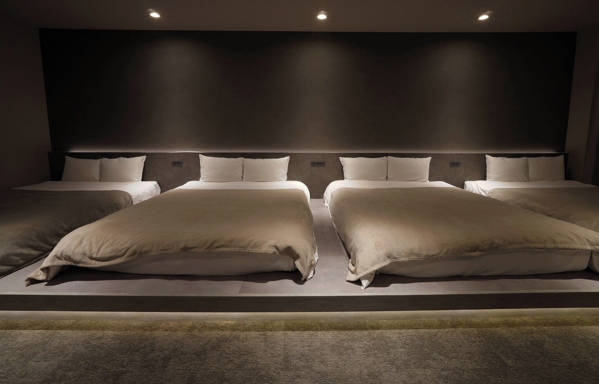 Ein Hotelzimmer mit vier nebeneinander stehenden Betten, einem dunklen Kopfteil und eingelassener Deckenbeleuchtung.