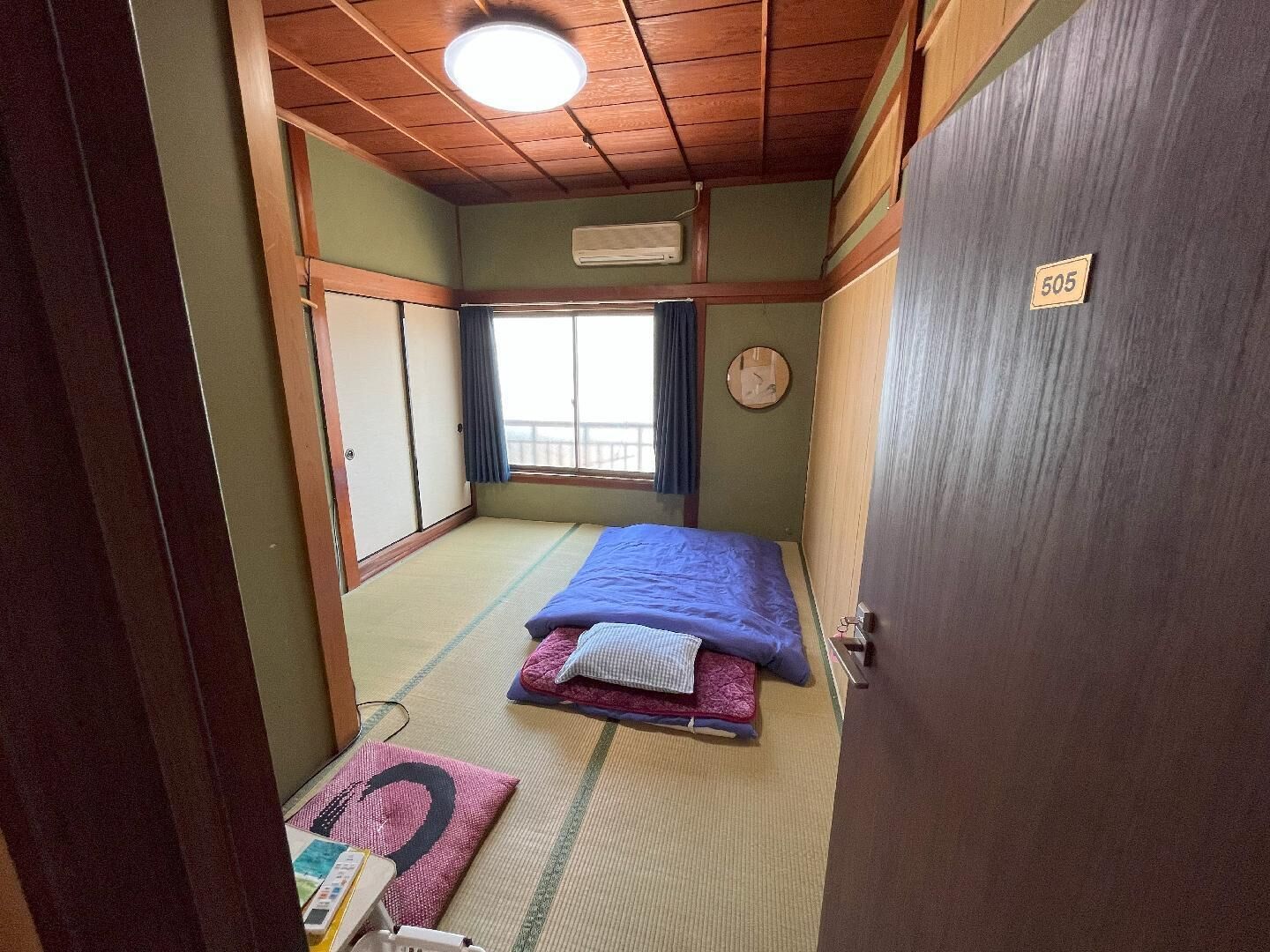 Ein traditioneller japanischer Raum mit Tatami-Böden, einem Bett mit blauer Decke, einer Holztür mit einer Nummer und einem Fenster mit Vorhängen.