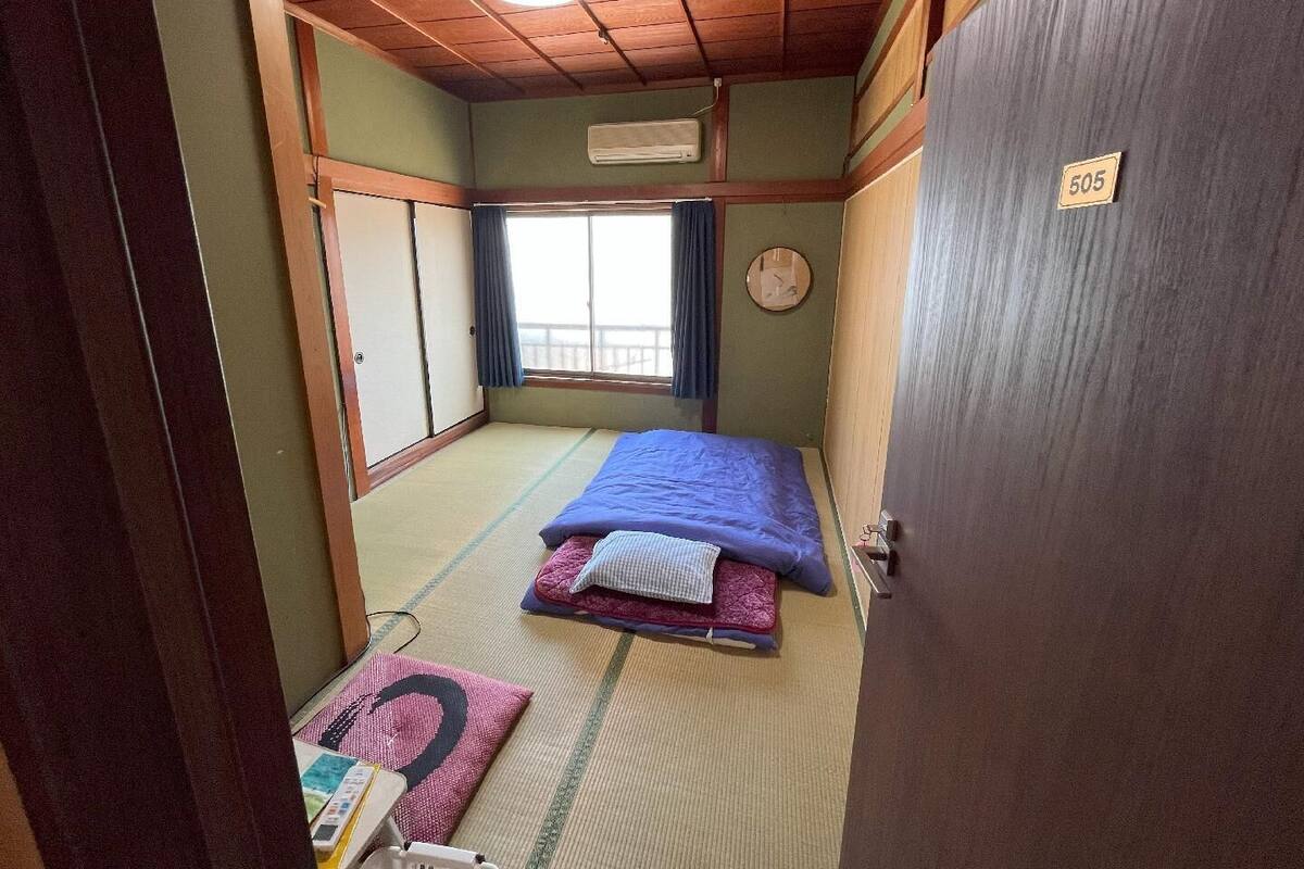 A traditional Japanese room with tatami flooring, a bed with a blue blanket, a wooden door with a number, and a window with curtains.