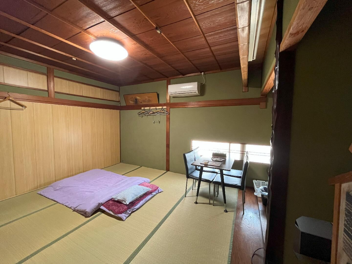 Ein Zimmer mit Tatami-Böden, einem Bett mit violetter Decke, einem Schreibtisch mit Stühlen und einem Fenster, das natürliches Licht hereinlässt.