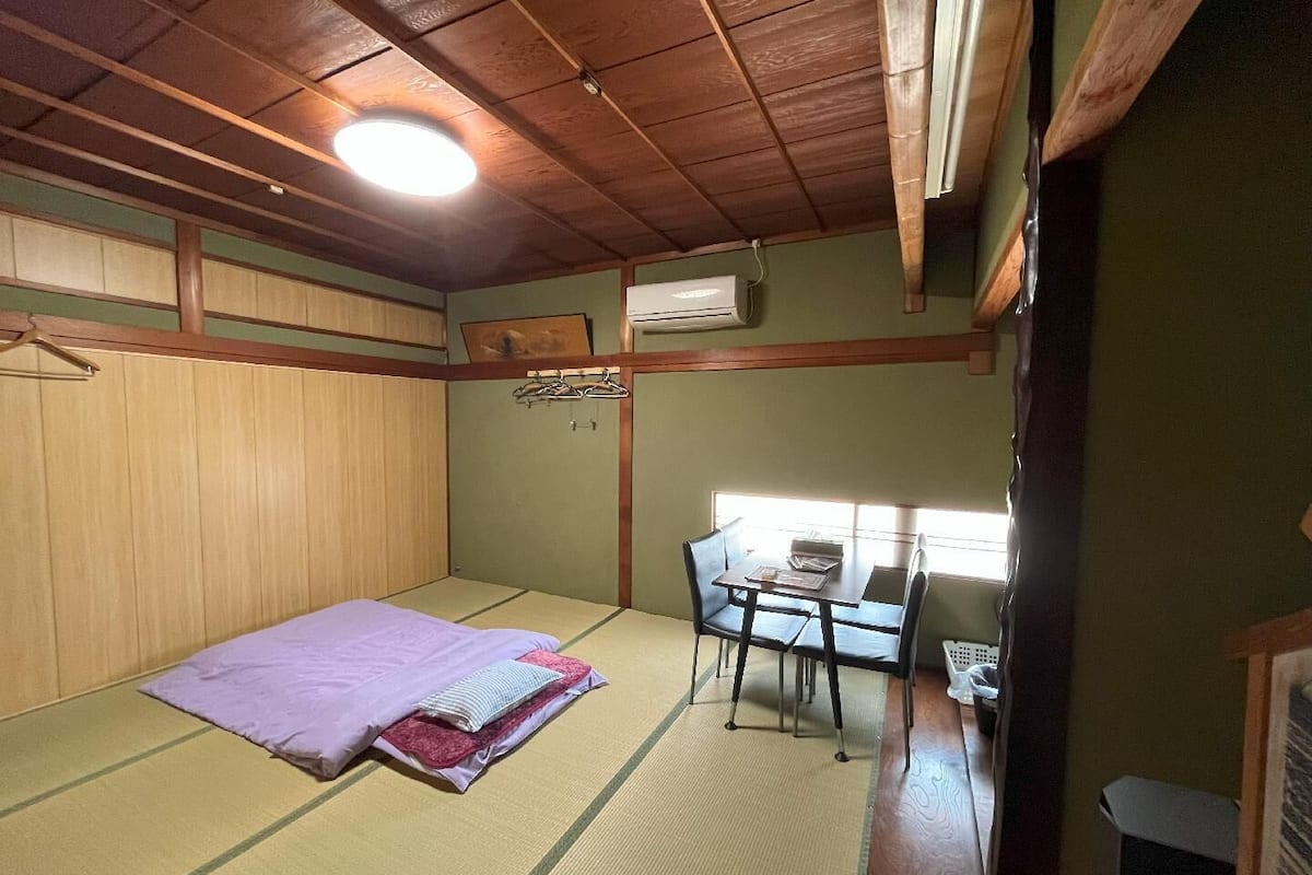 A room with tatami mat flooring, a bed with a purple blanket, a desk with chairs, and a window providing natural light.