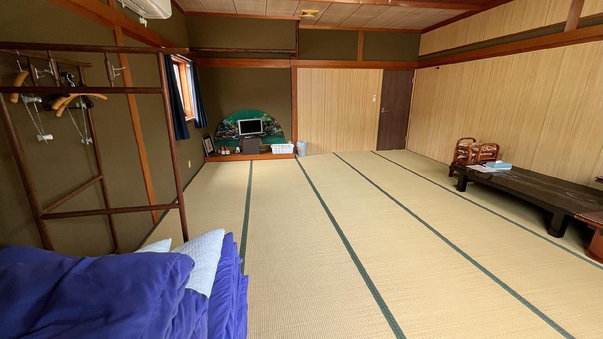Ein Raum mit Tatami-Matten, einem Futonbett, einem kleinen Tisch mit Gegenständen und einem Stuhl.