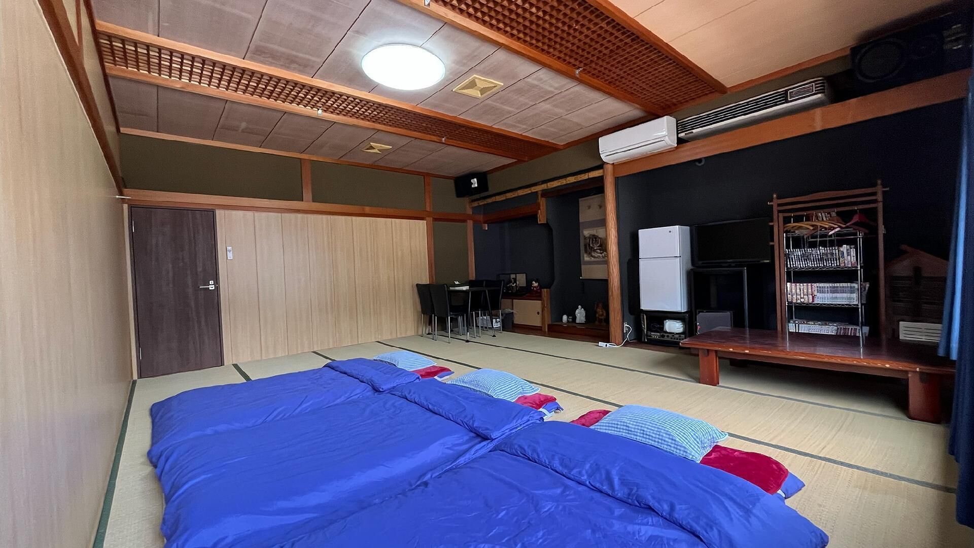Ein traditioneller japanischer Raum mit Tatami-Böden, einem Futon, einem Schreibtisch mit Stuhl, einem Fernseher und einem Bücherregal.