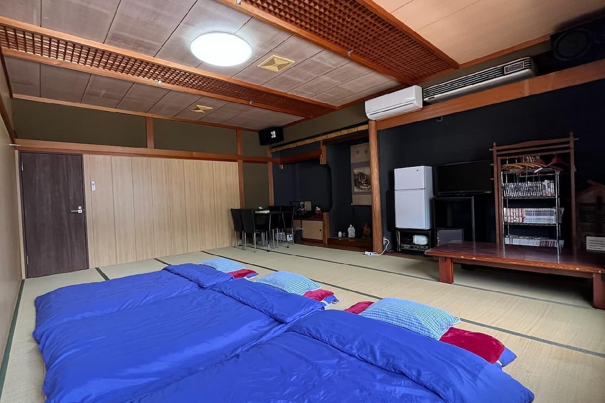 A traditional Japanese-style room with tatami flooring, a futon bed, a desk with a chair, a television, and a bookshelf.