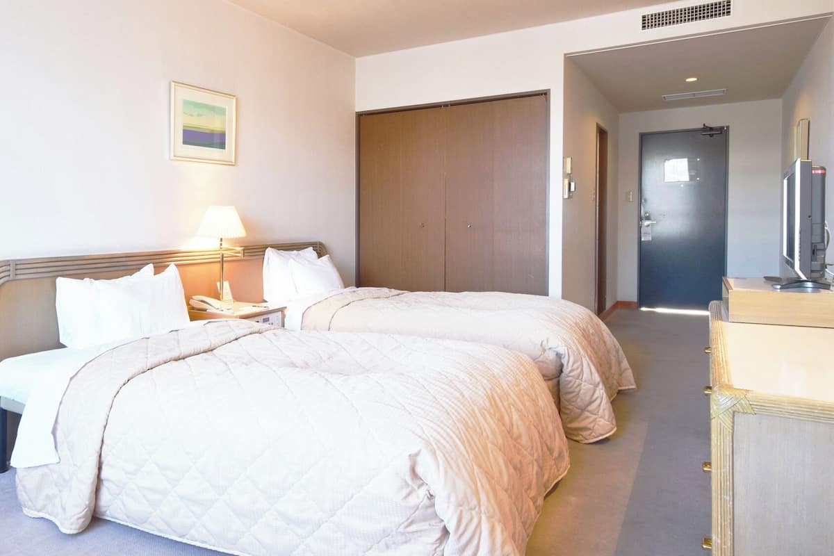A hotel room with two beds, a bedside table with a lamp, a framed picture on the wall, and a door leading to another room.