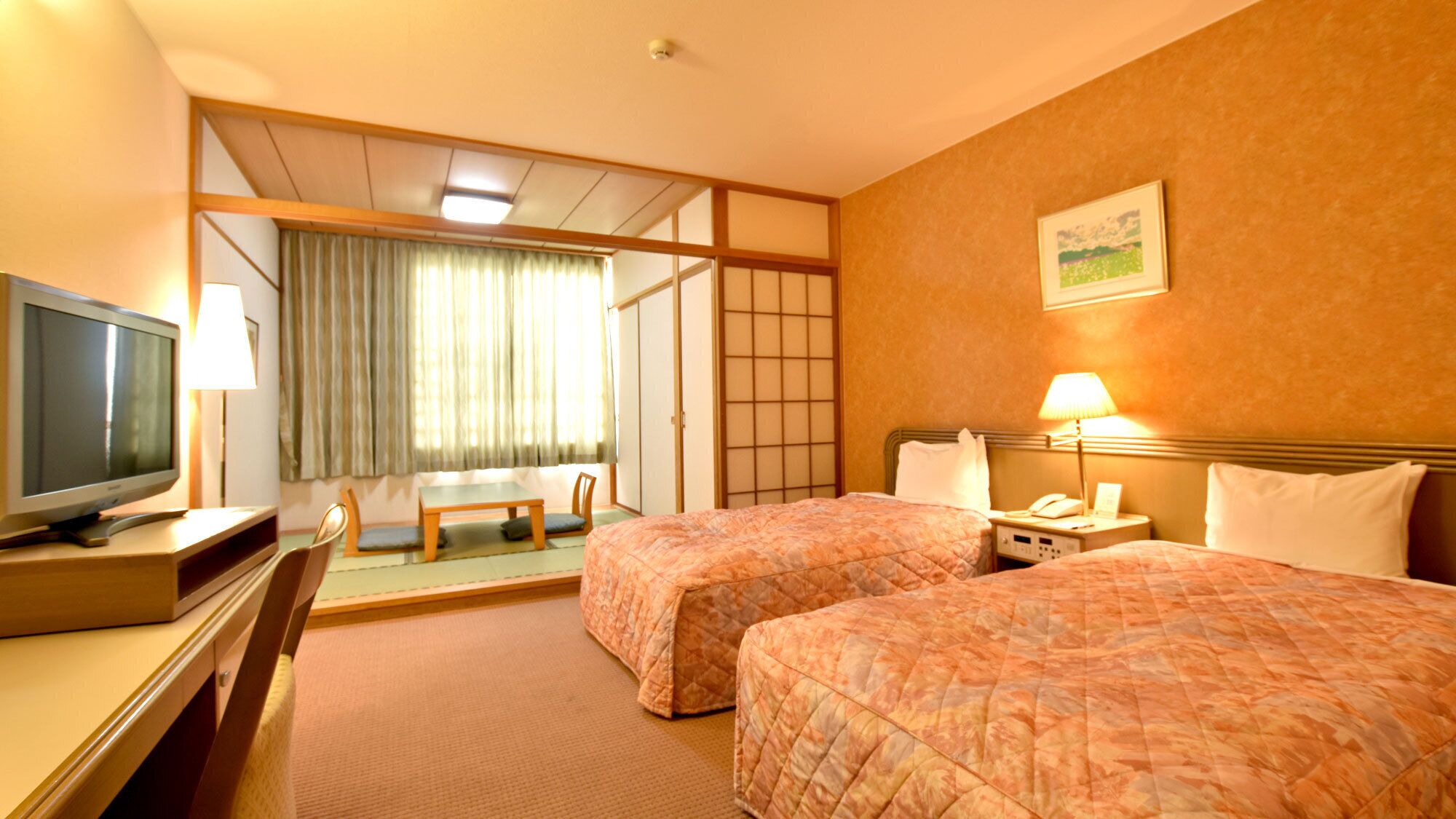 A hotel room with two beds, a desk with a television, a chair, a table, and a sliding door leading to a balcony.