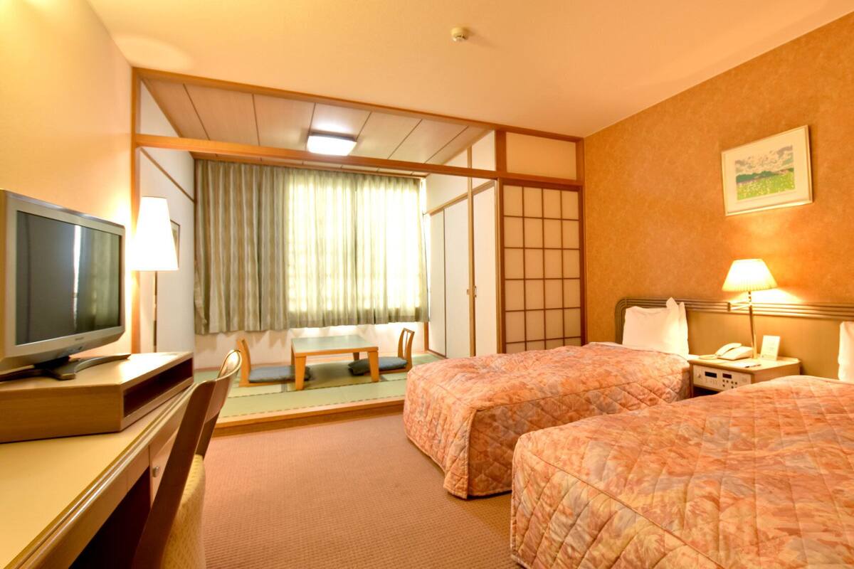 A hotel room with two beds, a desk with a television, a chair, a table, and a sliding door leading to a balcony.