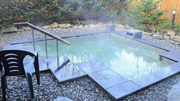 Una bañera de hidromasaje rectangular con cerramiento de vidrio, rodeada de guijarros y rocas, con una silla de madera y un banco de metal.