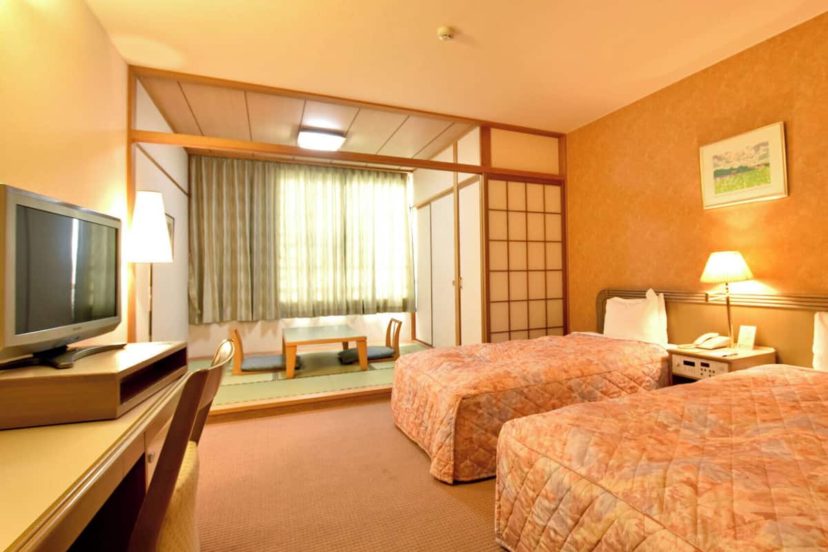 A hotel room with two beds, a desk with a television, a chair, a table, and a window with curtains.