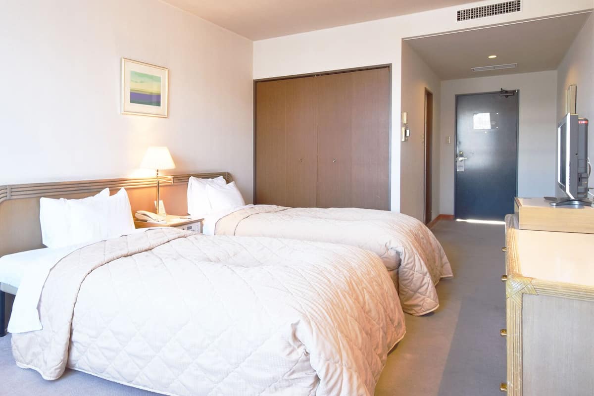 A hotel room with two beds, a bedside table with a lamp, a wardrobe, and a desk with a computer.