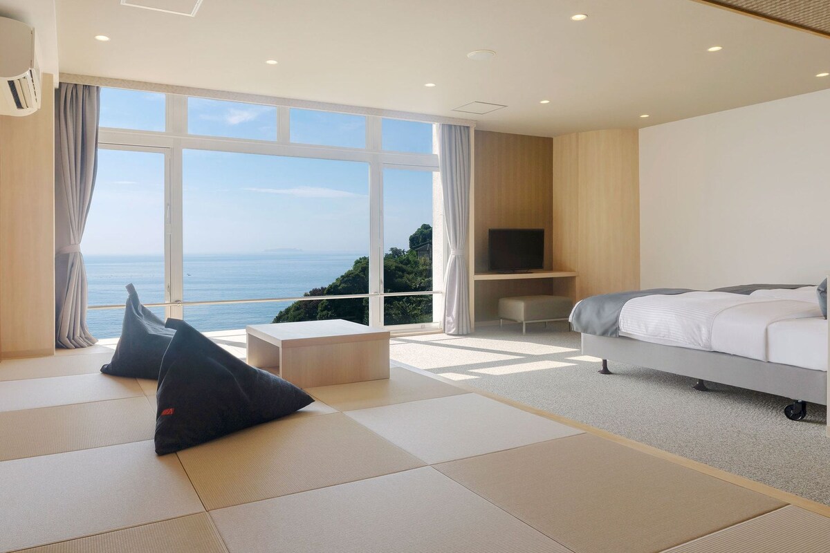 A hotel room with a bed, a TV, a seating area with cushions, and a large window offering a view of the sea.
