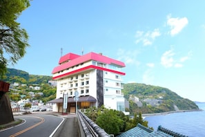 A multi-story hotel with a red roof, located near a coastal area with a view of the sea and hills. - Hotel Suiyotei (Atami)