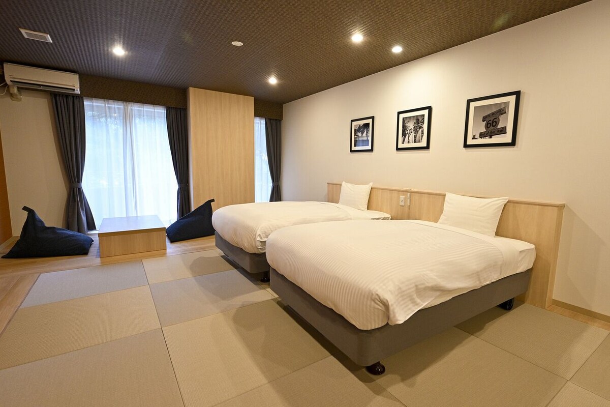 A hotel room with two beds, a large window with curtains, a wall-mounted air conditioner, and three framed pictures.