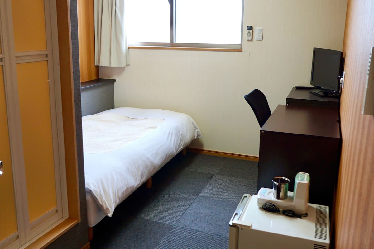 Habitación de hotel con cama, escritorio con computadora, silla, ventana con cortinas y termostato en la pared.