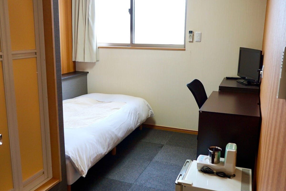Habitación de hotel con cama, escritorio con computadora, ventana con cortinas y un pequeño calefactor en el suelo.