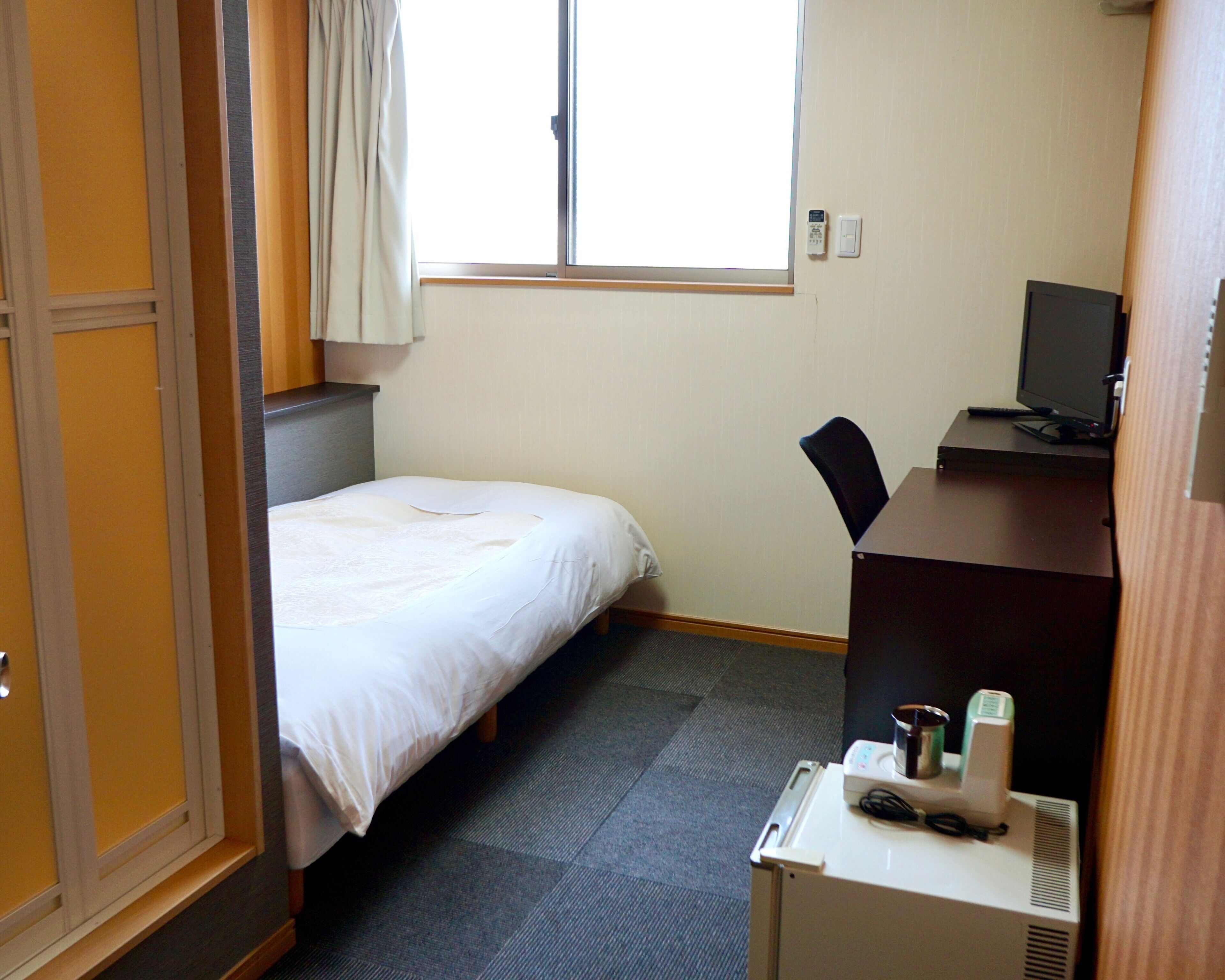 Habitación de hotel con cama, escritorio con computadora, silla, ventana con cortinas y termostato en la pared.