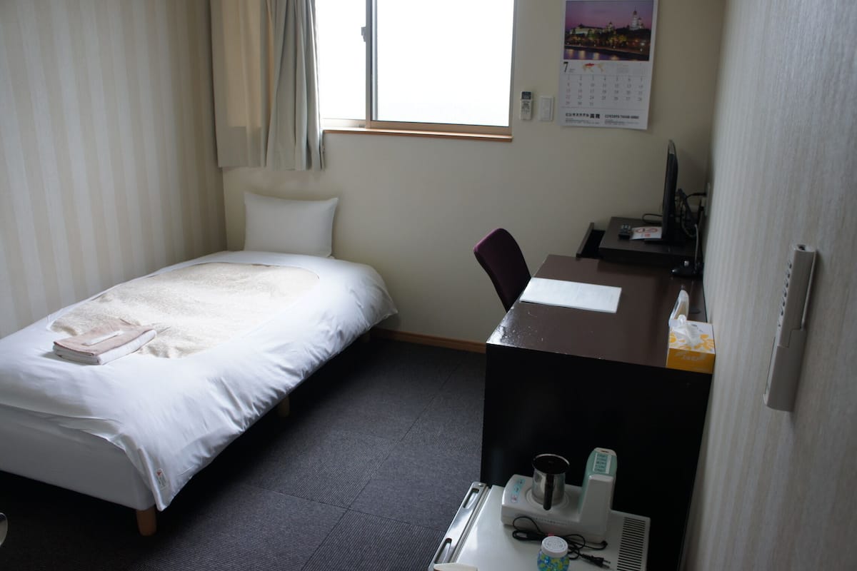 Una habitación de hotel con cama, escritorio, computadora, silla y ventana.