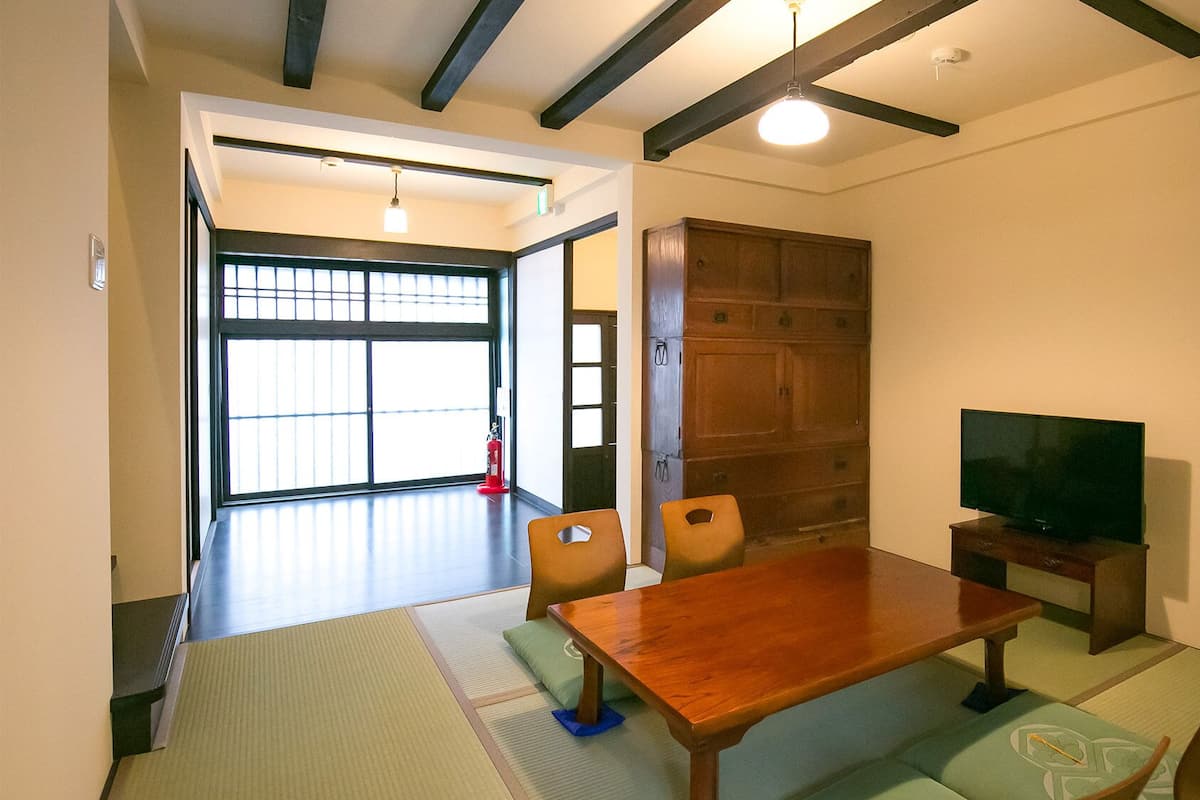 Ein Zimmer mit Tatami-Matten, einem Holztisch, Stühlen, einem Fernseher und einer Schiebetür zu einem weiteren Raum.