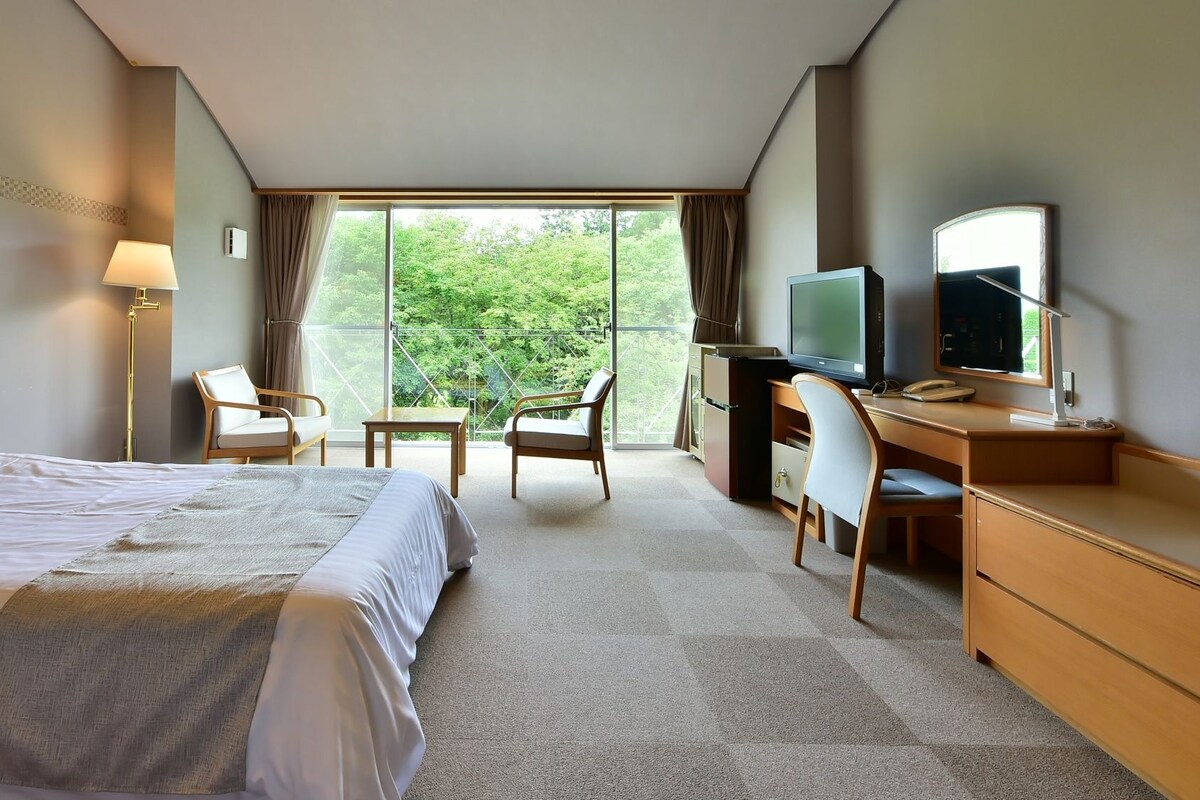 Habitación de hotel con cama, dos sillones, escritorio, televisor y un ventanal con vistas a zonas verdes.