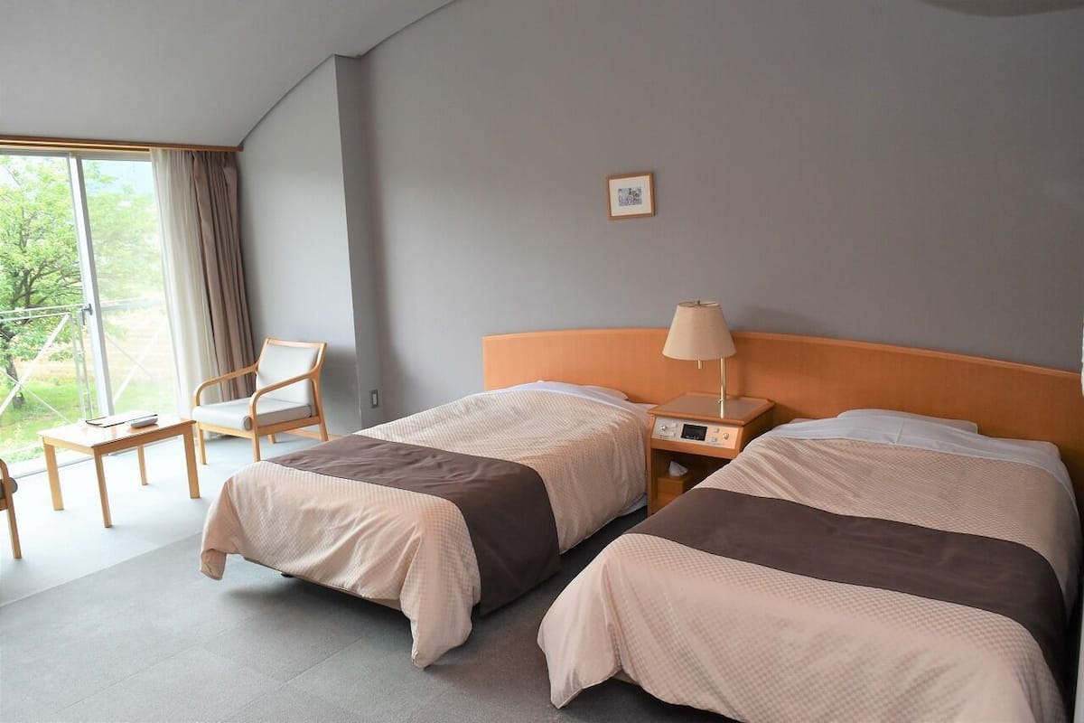 A hotel room with two beds, a chair, a table, a lamp, and a view of greenery.