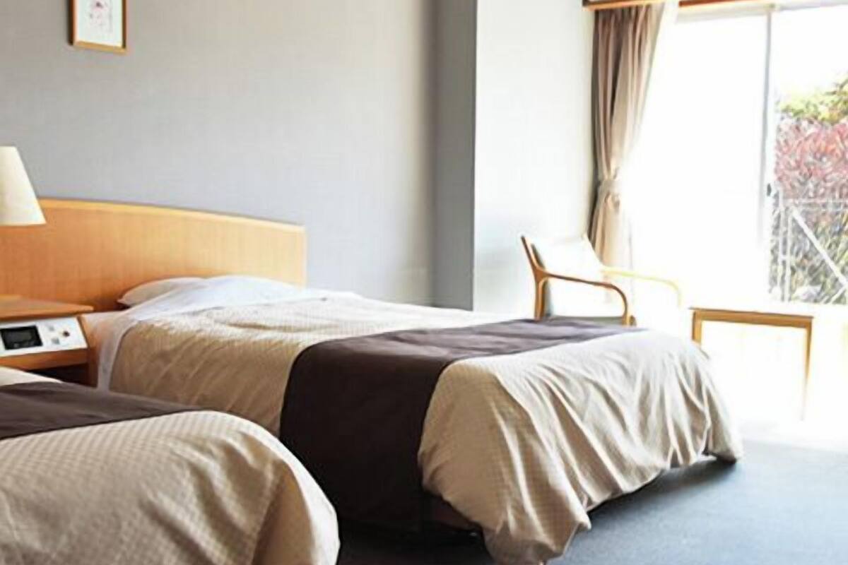 A hotel room with two beds, a bedside table with a lamp, a chair, and a large window with curtains.