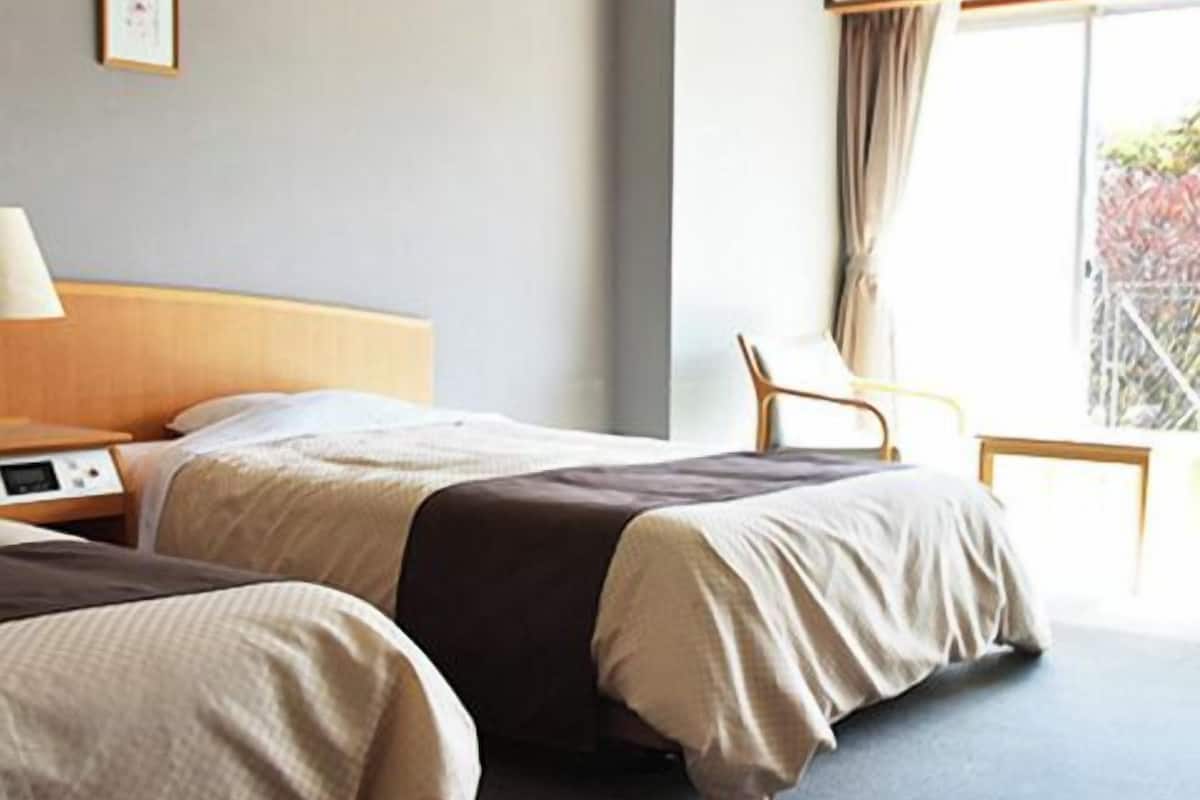 A hotel room with two beds, a bedside table with a lamp, a chair, and a large window with curtains.