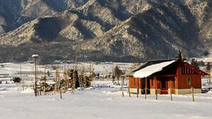 A wooden cabin with a snow-covered roof, surrounded by a snowy landscape and mountains in the background. - Suzumushiso (Matsukawa)