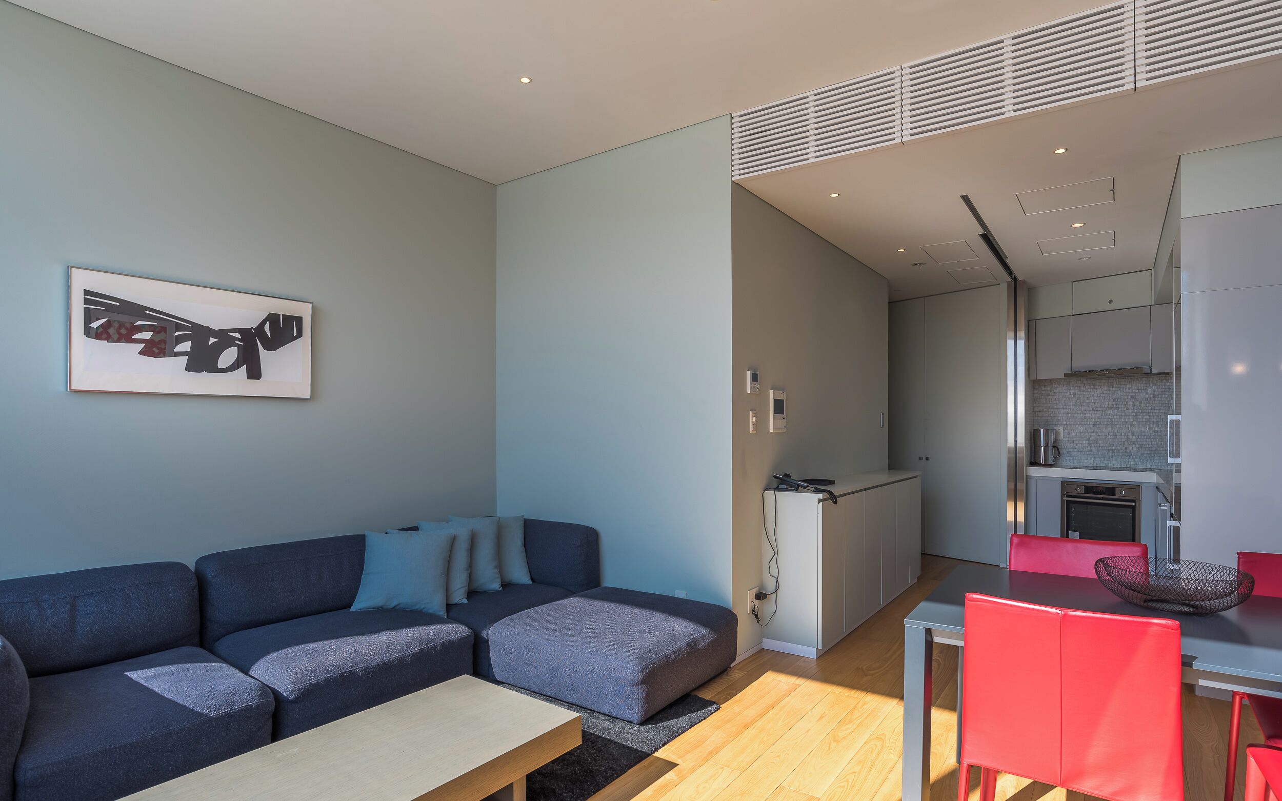 Un salon moderne avec un canapé, une table à manger entourée de chaises rouges, un coin cuisine et une œuvre d’art encadrée accrochée au mur.