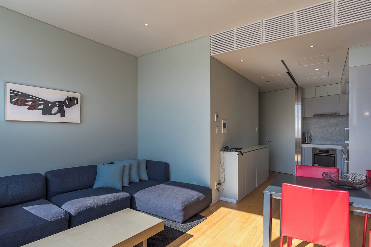 A modern living room with a sofa, a dining table with red chairs, a kitchen area, and a framed artwork on the wall.